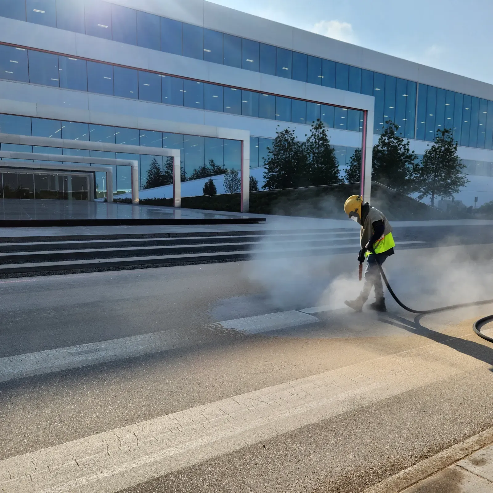 Worker sandblasting pavement outside a glass office building at sunrise