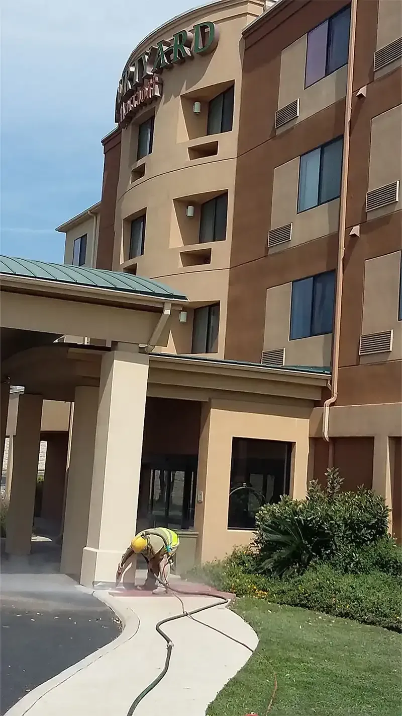 Technician sandblasting a concrete sidewalk outside a multi-story hotel