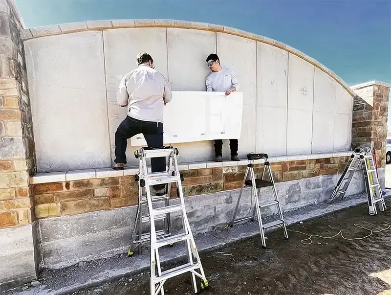 Two crew members on ladders resurfacing a stone entrance monument wall