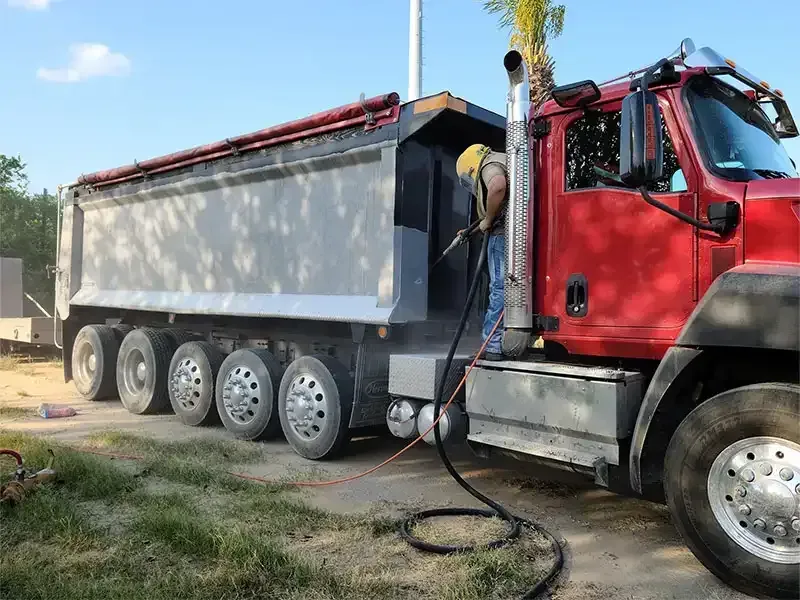 Worker sandblasting the steel bed of a red dump truck