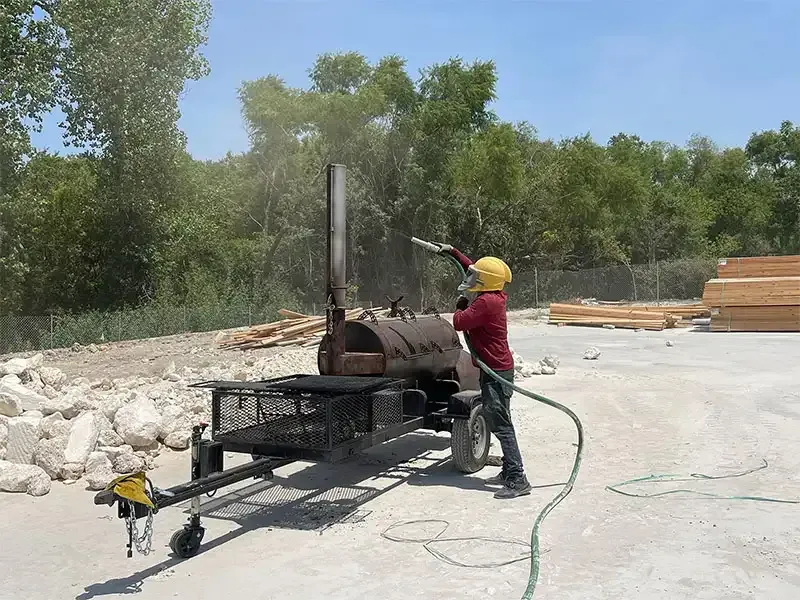 Technician blast cleaning a barbecue smoker trailer outdoors