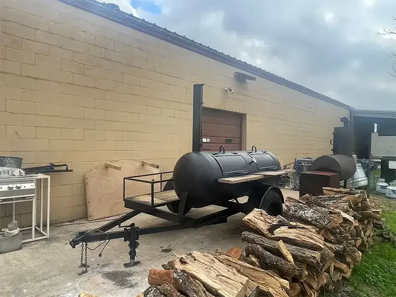 Black smoker trailer beside stacked firewood at a commercial kitchen