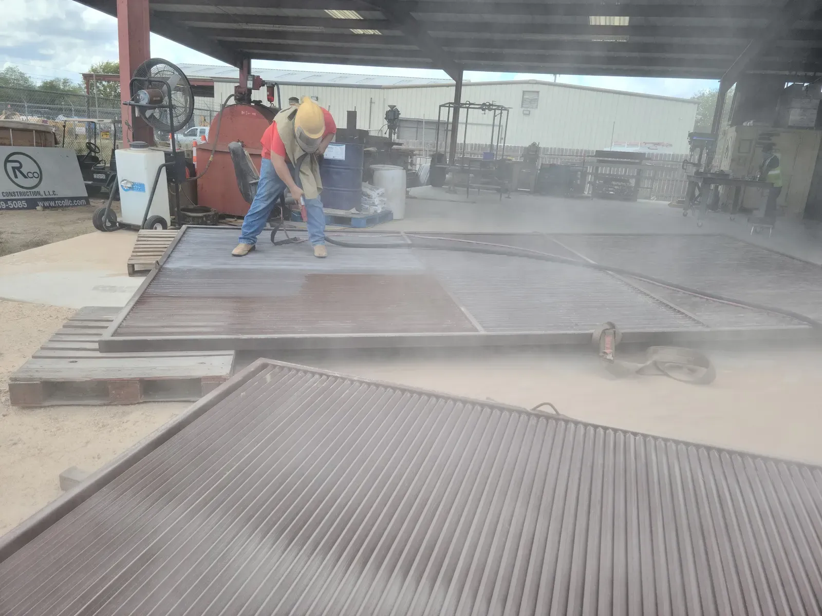 Worker sandblasting steel panels in an open-air fabrication shop