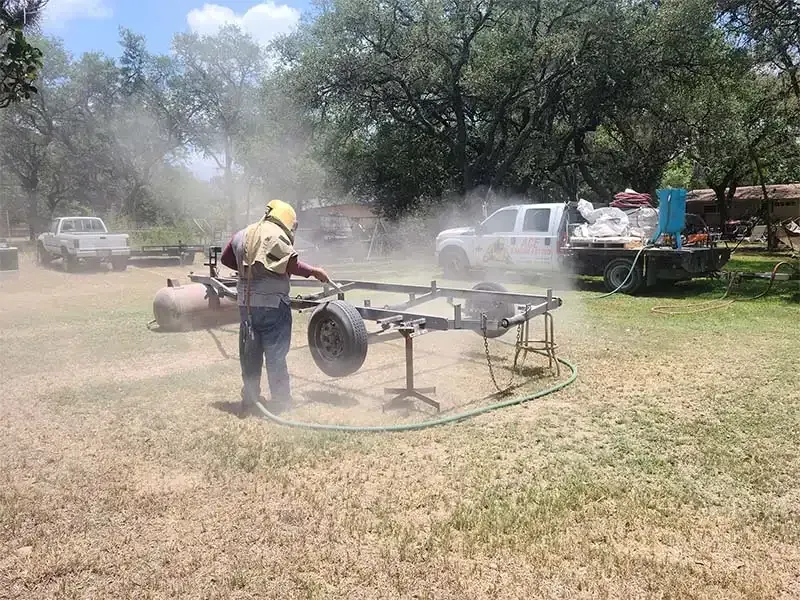 Worker sandblasting a steel trailer frame in a dusty field