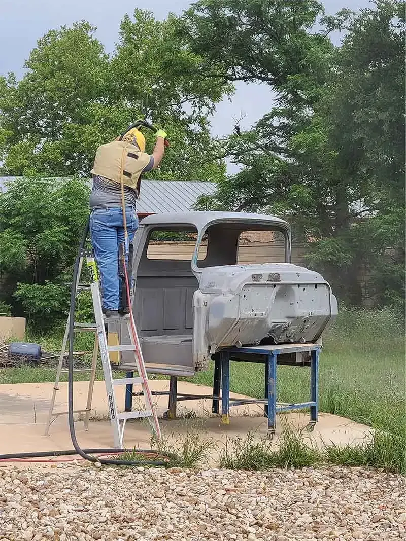 Technician on a ladder sandblasting a vintage pickup truck cab